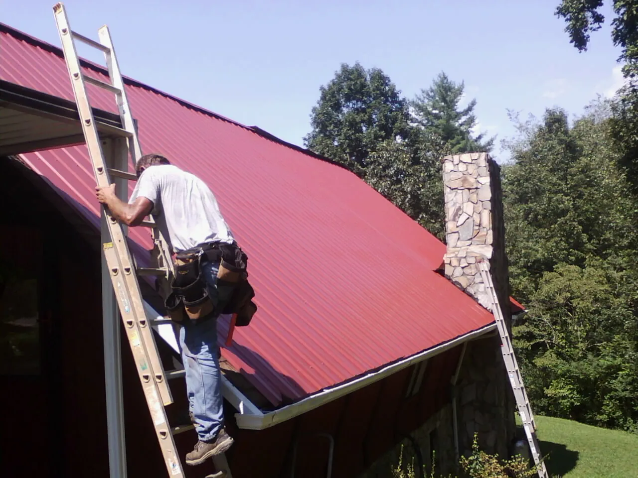 Roofer on a ladder beside a red metal roof and stone chimney — Soffit & Fascia Repair by Roofing Brothers in Atlasburg